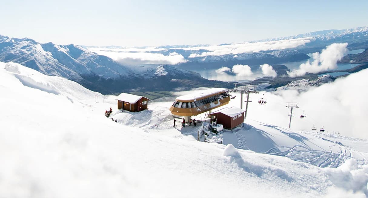 A chairlift station at Treble Cone Ski Field sits atop a snowy mountain. In the background, stunning views of Lake Wanaka and snow-covered peaks stretch into the distance under a clear sky.