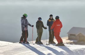 A ski instructor in a red jacket talks with three skiers on a snowy slope. The morning light creates long shadows, with a mountain lodge visible in the background under a serene sky.