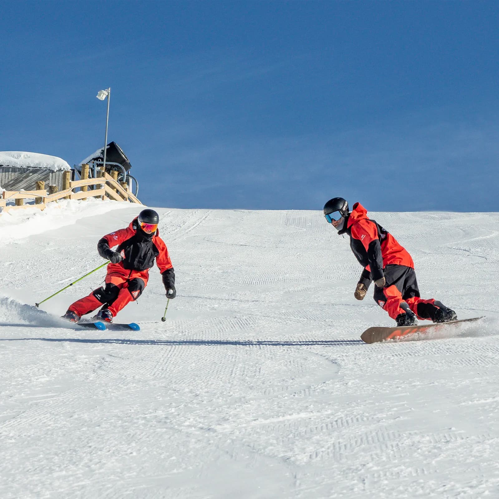 A skier and a snowboarder side by side on a snowy slope. Both are dressed in matching red and black winter sportswear. The skier, using poles, carves elegantly through the snow, while the snowboarder, appearing in a parallel pose, glides smoothly on their board. The background shows a clear blue sky and a small ski hut at the top of the slope.