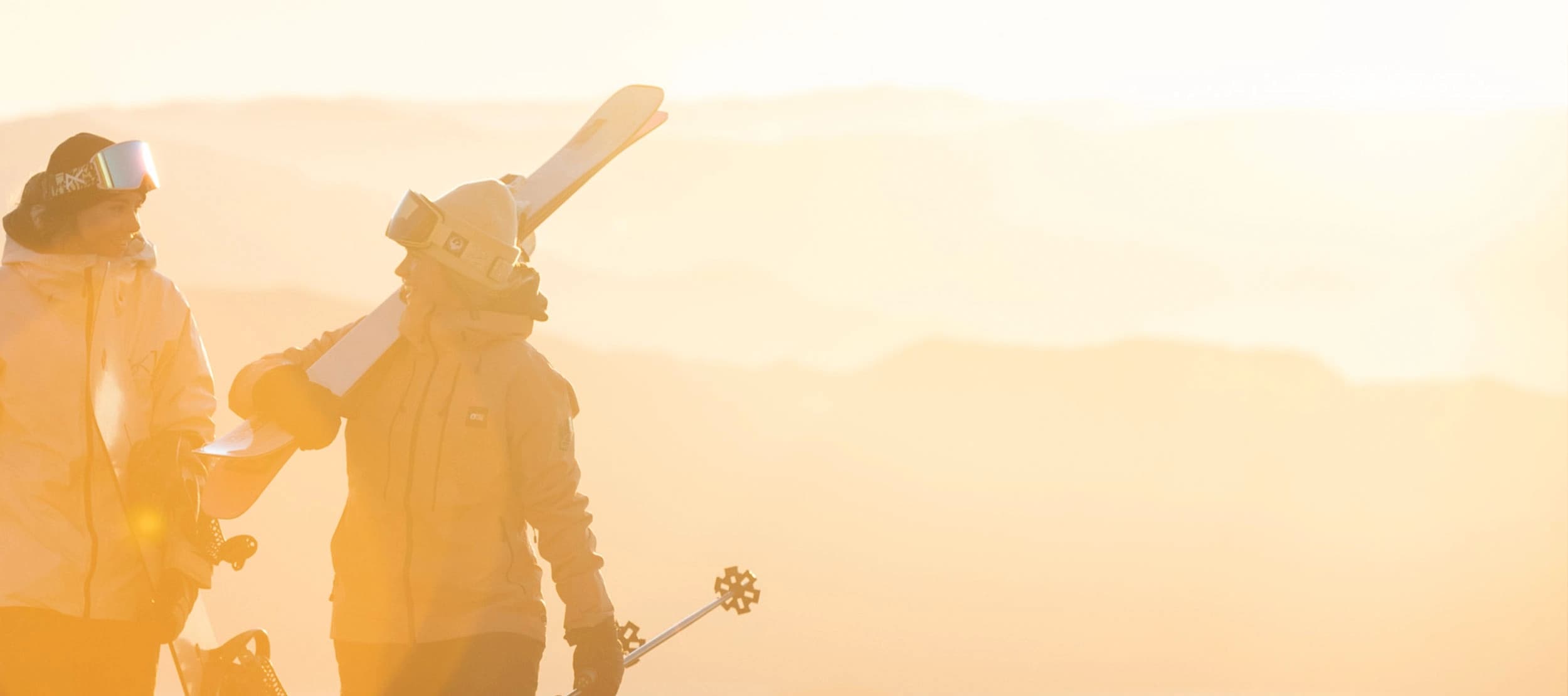 Two friends talk as they carry their ski and snowboard gear towards the bottom of the ski chairlift as the sunrises behind them.