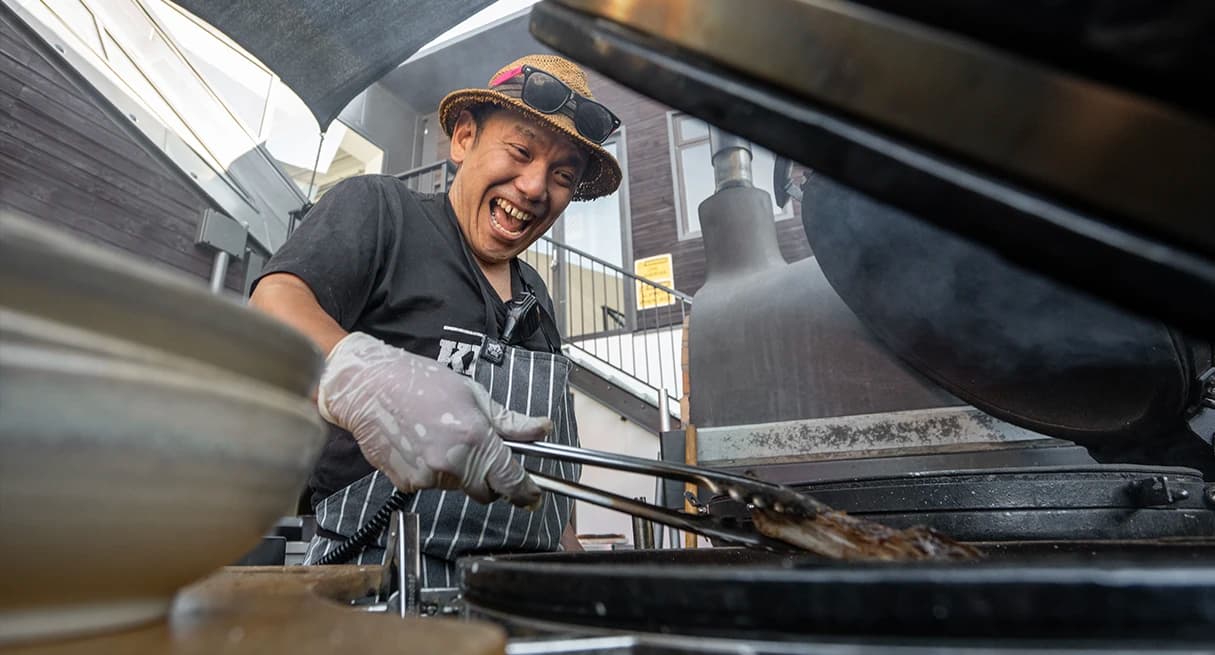 A smiling chef in a striped apron grills food at an outdoor kitchen, holding tongs and working over a hot grill, enjoying the sunny day.