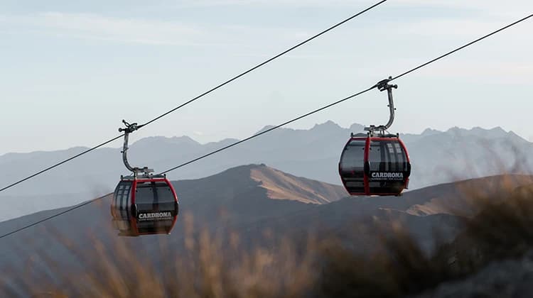 Two red gondolas glide along cables above a rugged alpine landscape at Cardrona Alpine Resort in New Zealand. The distant mountain ranges create a scenic backdrop under a clear sky.