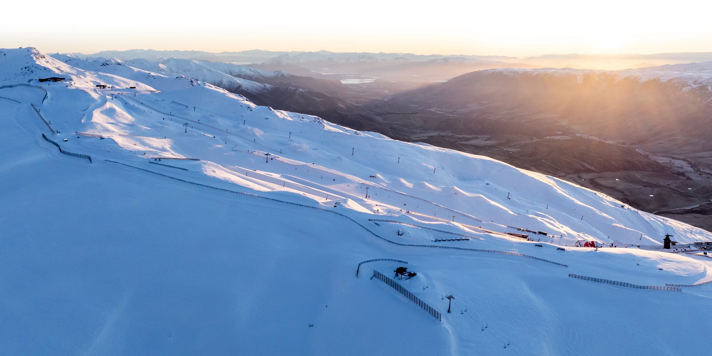 An expansive view of a snow-covered ski resort at sunrise, with soft light illuminating the slopes and surrounding mountain landscape.