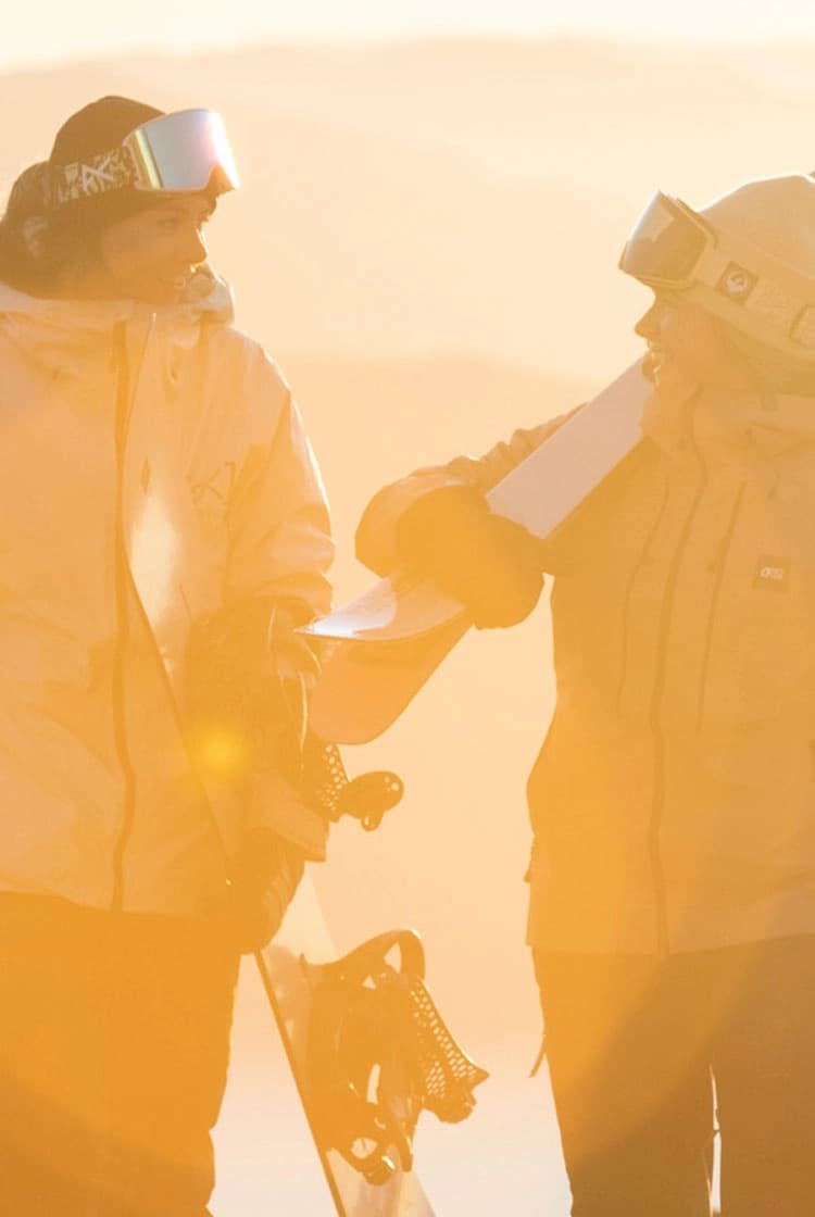 Two friends talk as they carry their ski and snowboard gear towards the bottom of the ski chairlift as the sunrises behind them.