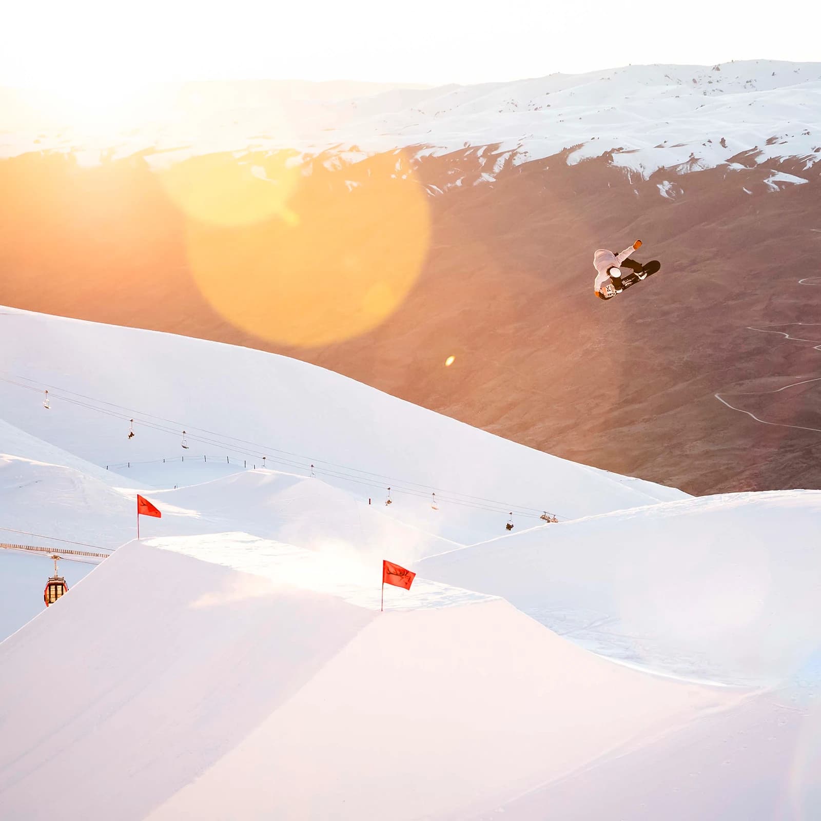 A snowboarder performs a high jump trick over a snow-covered mountain terrain, with ski lifts in the background and the sun casting a warm glow over the scene.