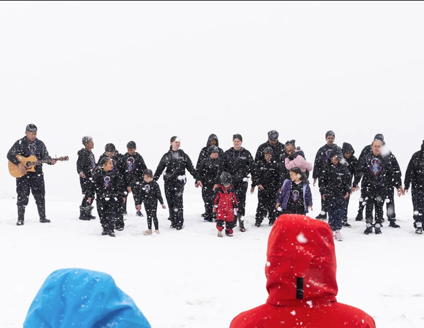 A group of people perform a haka in the snow while two guitarists play, as an audience in colorful jackets watches. Snow falls gently around them, creating a serene winter scene.