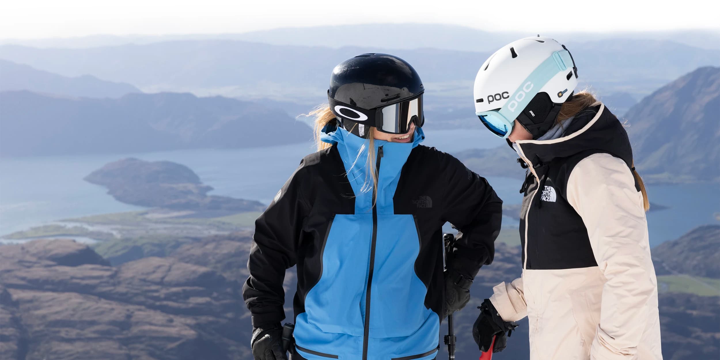 Two skiers wearing helmets and goggles stand on a snowy mountain, chatting with a scenic view of a lake and mountains in the background.