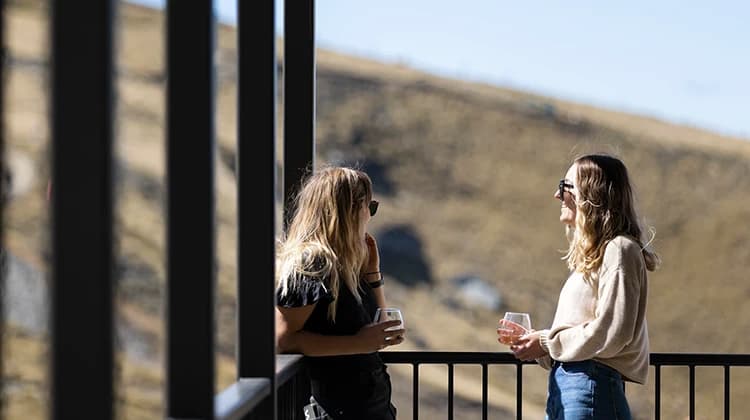 Two women enjoy a drink while standing outside on a balcony on a blue-sky summer day.