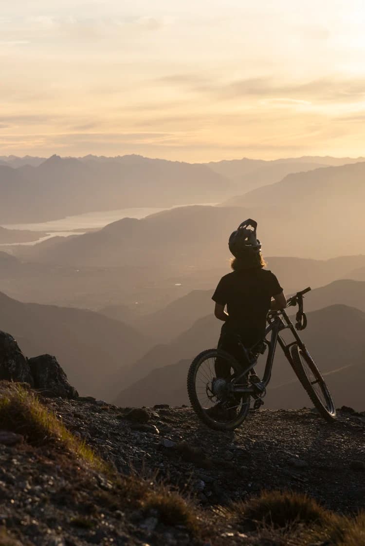 A mountain biker stands with their bike at sunset, overlooking the expansive Cardrona mountain range and valleys below.