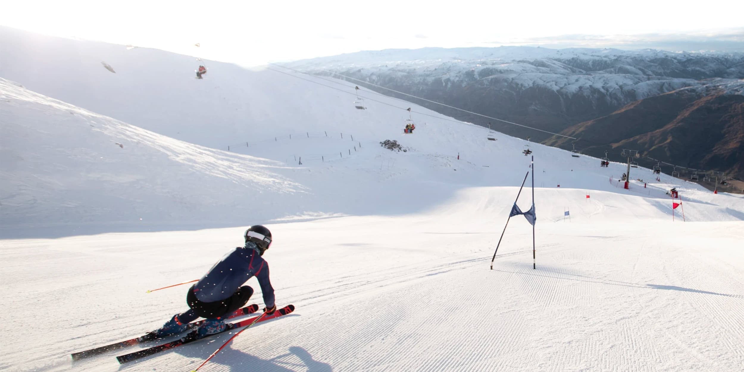 A skier in full gear speeding down a snow-covered slope, navigating between gates. The skier is in a low, aerodynamic position with ski poles extended behind them. The smooth, freshly groomed snow and a clear, expansive view of the mountain landscape with a ski lift in the background suggest a high-altitude ski area on a sunny day.