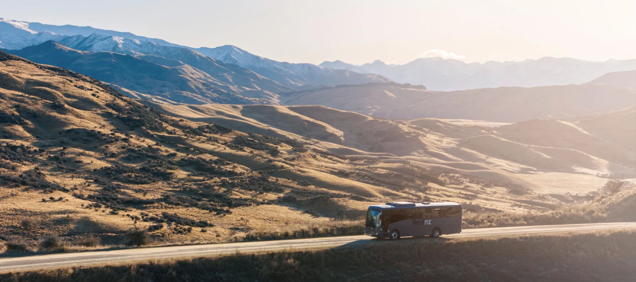 A tour bus travels along a winding road through a golden, mountainous landscape at sunset. Snow-capped peaks rise in the background, creating a serene and scenic vista.