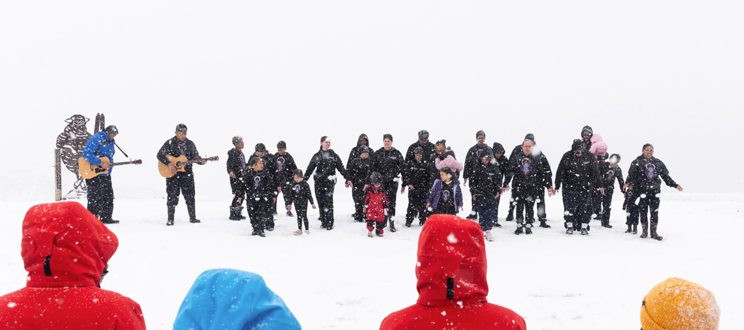 A group of people perform a haka in the snow while two guitarists play, as an audience in colorful jackets watches. Snow falls gently around them, creating a serene winter scene.