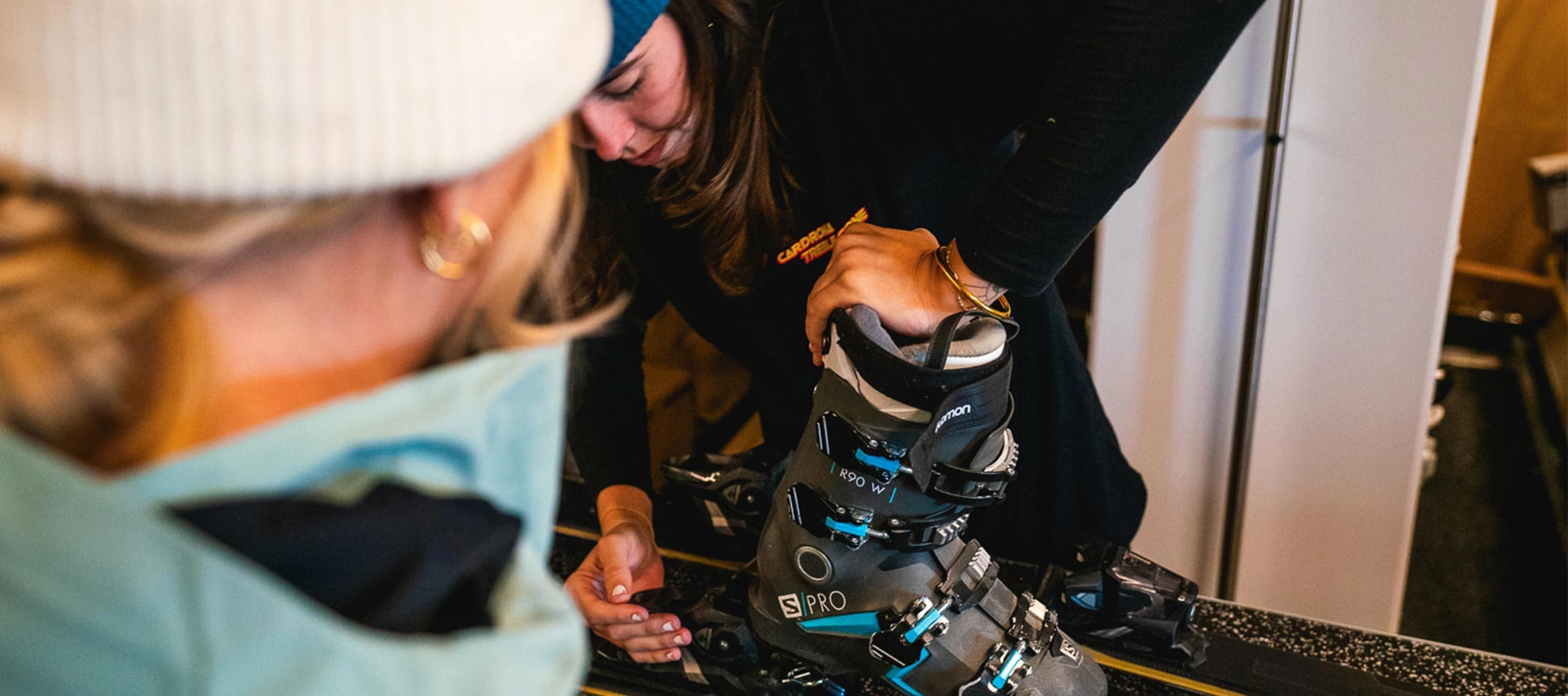 An indoor scene at a ski rental shop with a female technician adjusting ski boots for a customer. The technician, wearing a black long-sleeve shirt with the "Cardrona Rentals" logo, is focused on fitting a black and blue Salomon ski boot onto the bindings. The customer, partially visible, is wearing a light blue jacket and a white beanie.