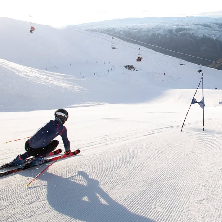 A skier in full gear speeding down a snow-covered slope, navigating between gates. The skier is in a low, aerodynamic position with ski poles extended behind them. The smooth, freshly groomed snow and a clear, expansive view of the mountain landscape with a ski lift in the background suggest a high-altitude ski area on a sunny day.