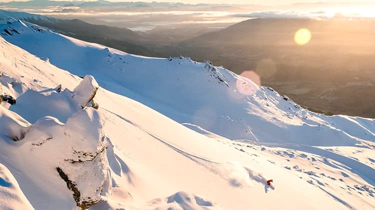 A lone skier in red glides down a vast, sunlit snow-covered mountain. The golden sunset and distant peaks create a serene backdrop.