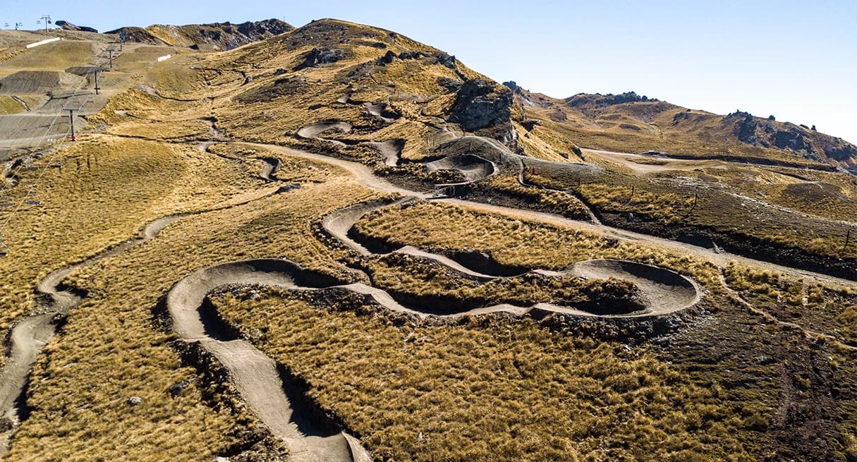 Aerial view of Cardrona Alpine Resort with winding dirt biking trails on golden-brown slopes, surrounded by rocky hills under clear skies.