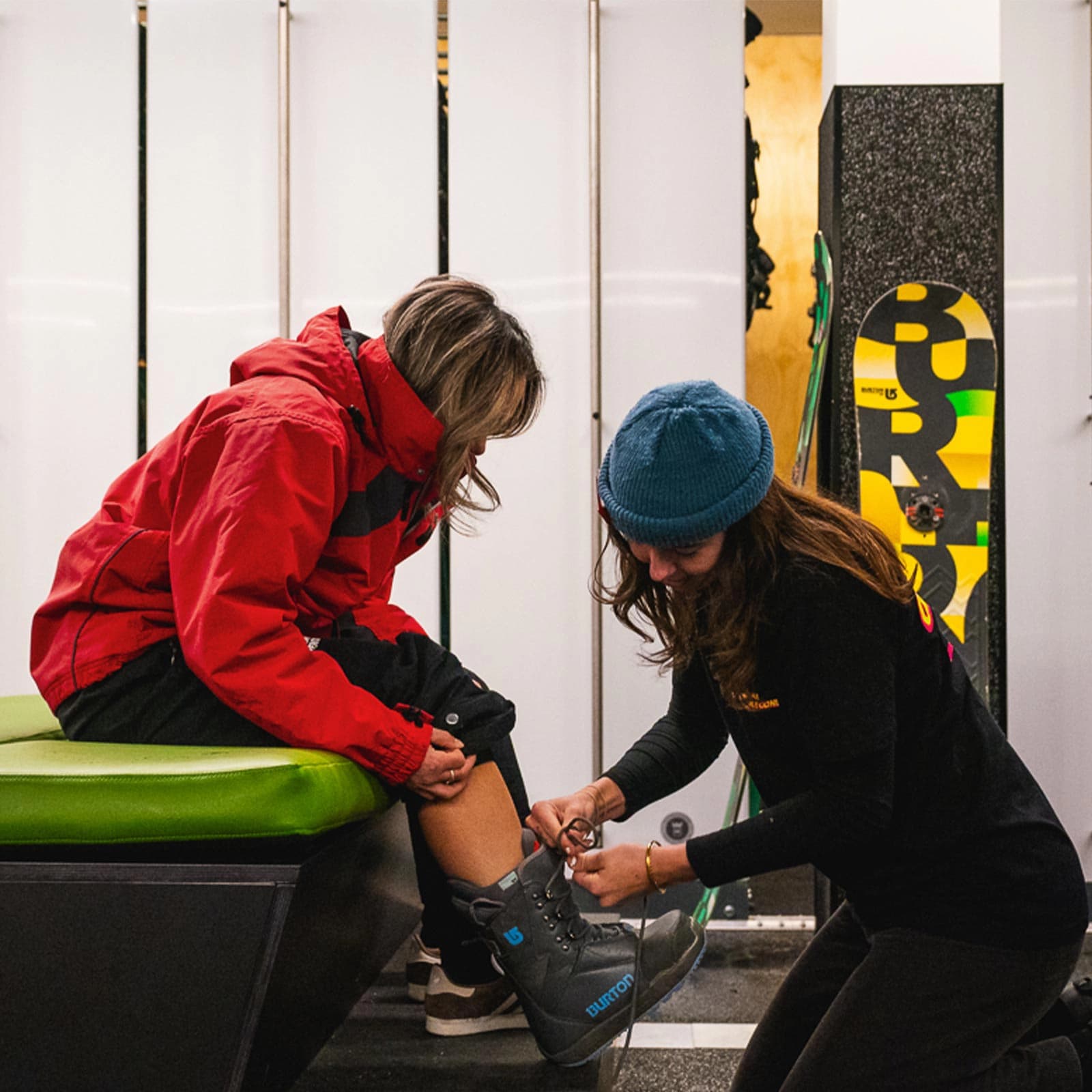 Inside a ski rental facility, a female technician in a black Cardrona Rentals uniform adjusts the straps of a grey Burton snowboard boot on a customer's foot. The customer, wearing a red jacket and seated on a green cushioned bench, watches the fitting process. In the background, snowboards and ski equipment are neatly stored in white lockers.