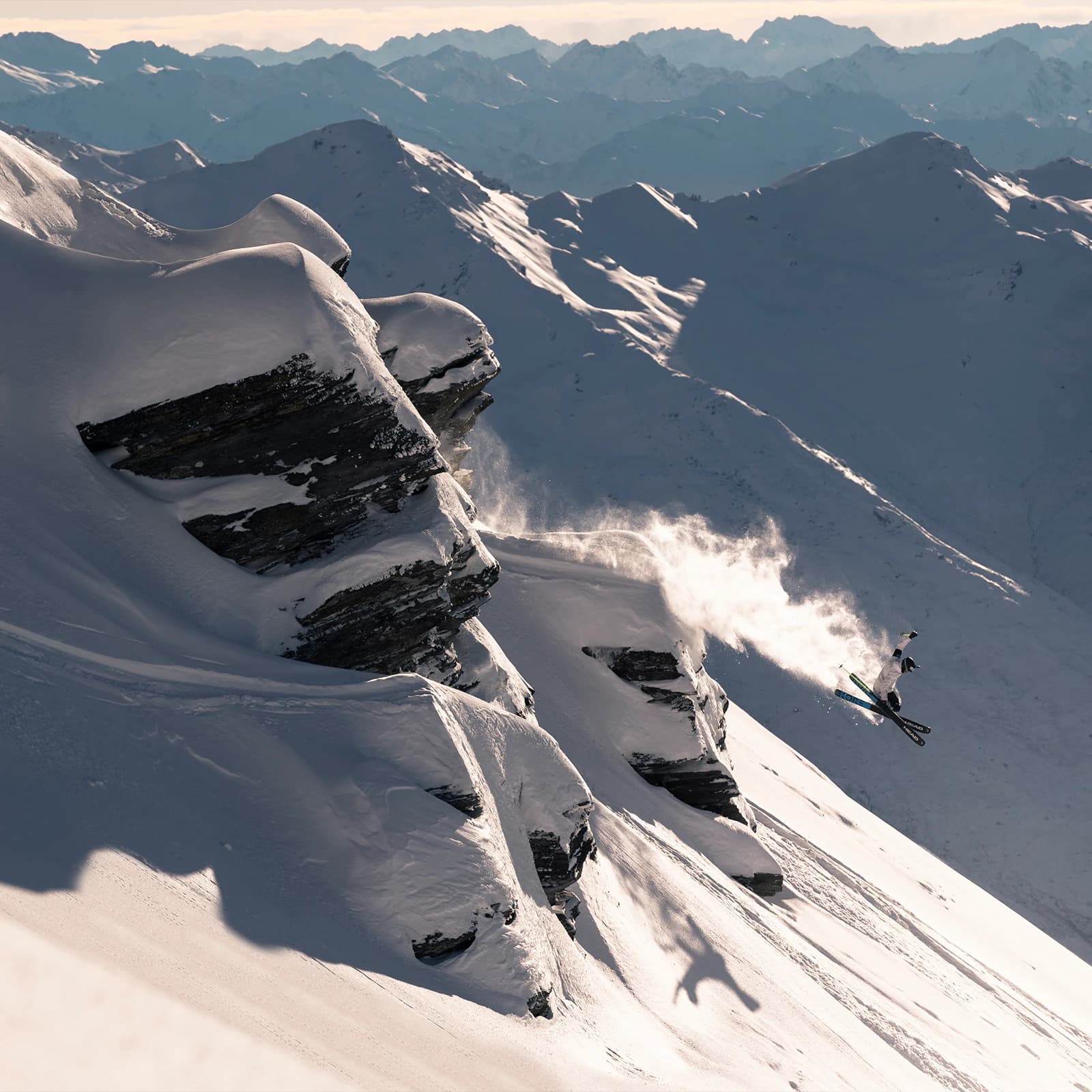 A skier launches off a rugged, snow-covered cliff, leaving a trail of powder in the air, with layers of distant, shadowed mountains forming a dramatic backdrop.