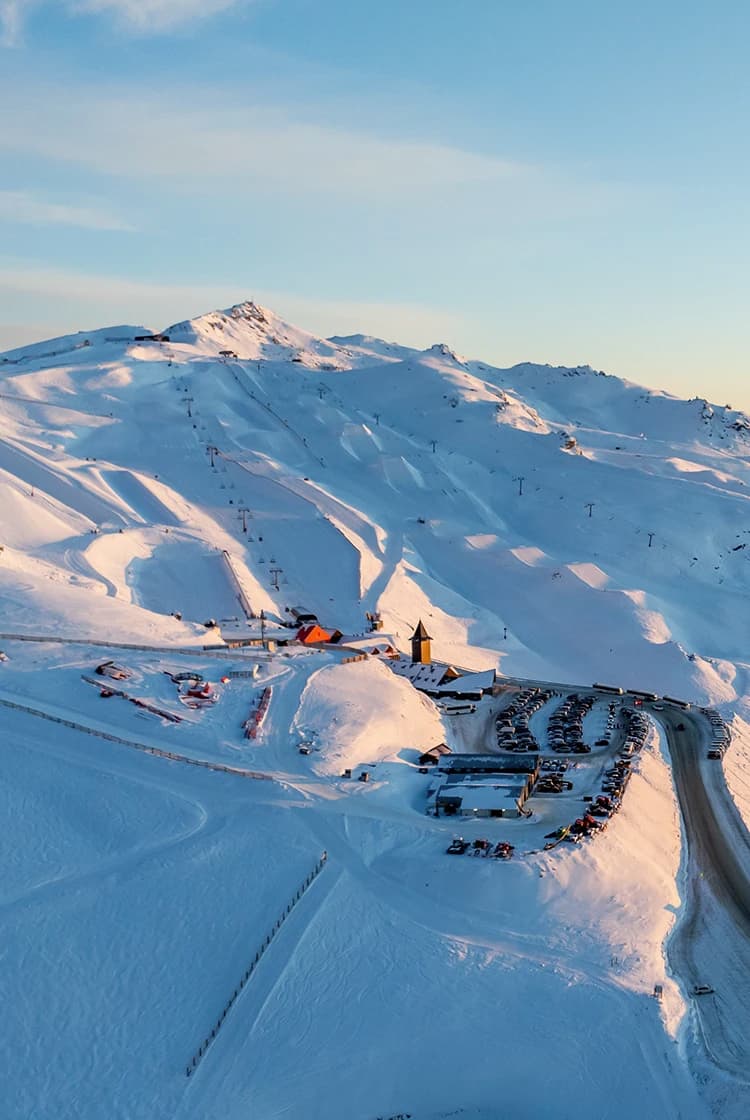 An aerial view of a snow-covered Cardrona Alpine Resort at sunset, featuring winding roads, ski lifts, and slopes illuminated by the soft light. The mountain peaks extend into the distance, creating a breathtaking winter landscape.