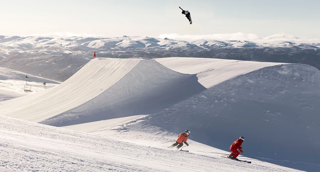 Two skiers descend a slope at Cardrona Alpine Resort, while a snowboarder performs an aerial trick above a large snow ramp, with mountains in the background.