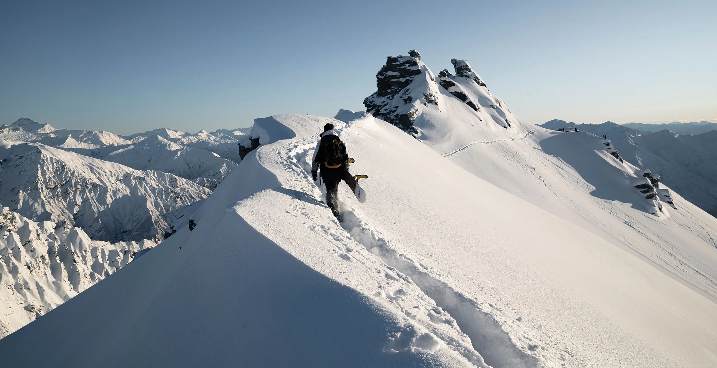 A snowboarder hikes along a narrow, snow-covered mountain ridge with breathtaking views of rugged peaks and deep valleys in the distance under a clear blue sky.
