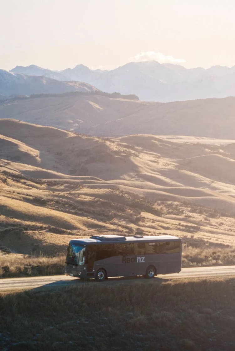 A tour bus travels along a winding road through a golden, mountainous landscape at sunset. Snow-capped peaks rise in the background, creating a serene and scenic vista.