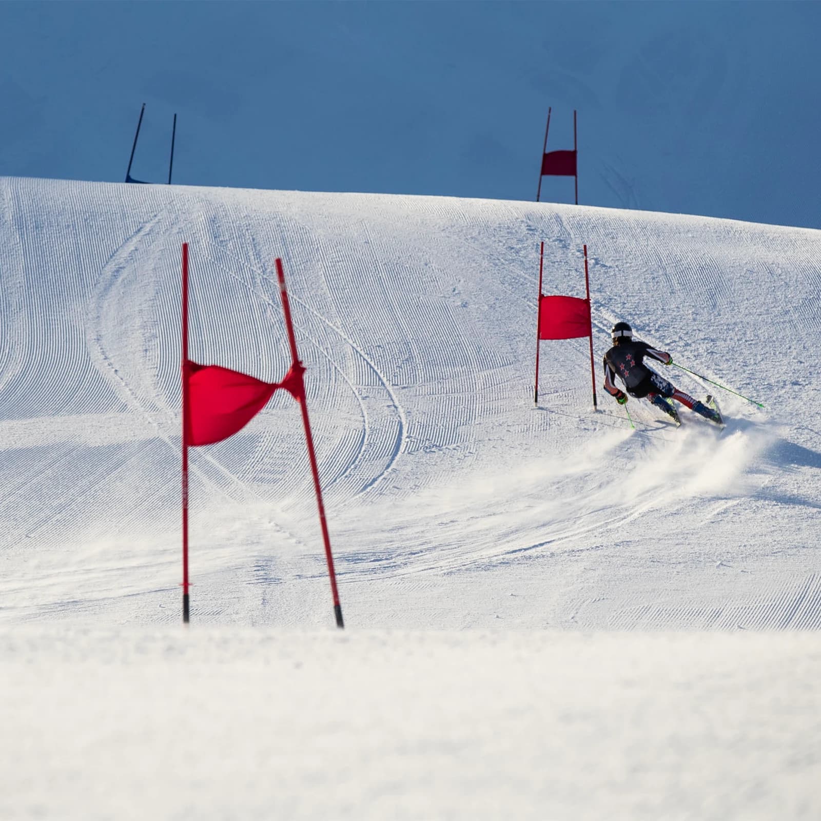 A skier in mid-action during a slalom race, carving a sharp turn around a red gate on a well-groomed piste. The skier is dressed in a black racing suit with a helmet and goggles. The background features a series of red gates extending up a snowy slope, emphasising the challenge of the course. The pristine condition of the snow and the clear blue skies create a vivid and exhilarating winter sports scene.