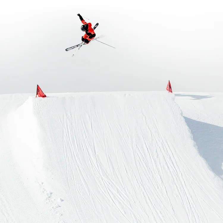 A skier performs a mid-air trick over a snowy slope, with red markers highlighting the edges of the jump, set against a bright, overcast sky.