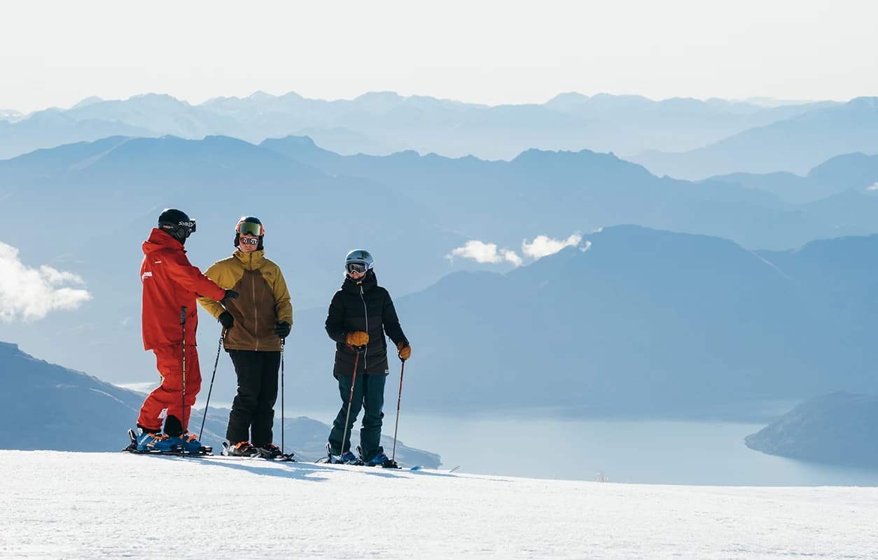 Three skiers stand on a snowy slope, engaged in conversation, with a breathtaking backdrop of distant mountains and a serene lake below.