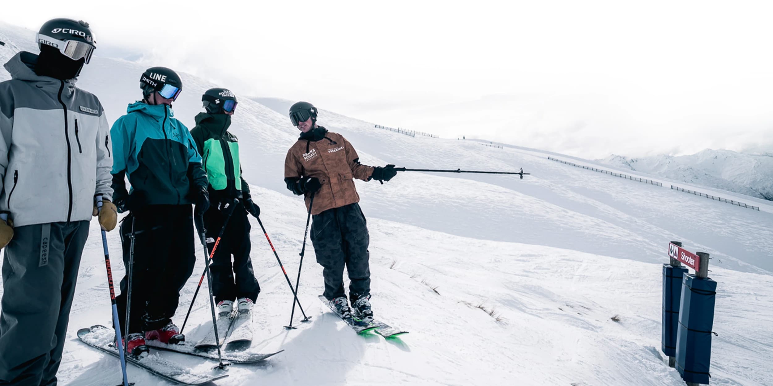 Four skiers on a snow-covered slope, dressed in winter ski gear including helmets and goggles. The skiers are equipped with poles and are standing on skis.