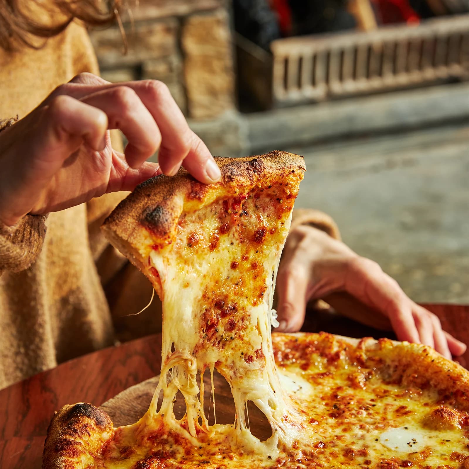 Close up of someone pulling away the first slice from a margherita pizza with a burning fire place in the background.