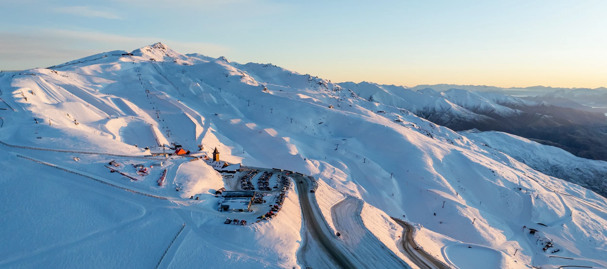 An aerial view of a snow-covered Cardrona Alpine Resort at sunset, featuring winding roads, ski lifts, and slopes illuminated by the soft light. The mountain peaks extend into the distance, creating a breathtaking winter landscape.