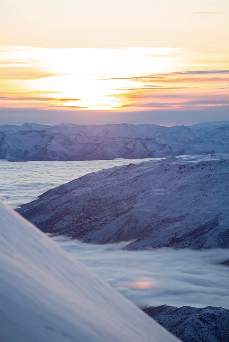 A serene mountain landscape at sunset, with the sun casting a warm glow over snow-covered peaks and a sea of clouds filling the valleys below, creating a peaceful and majestic scene.