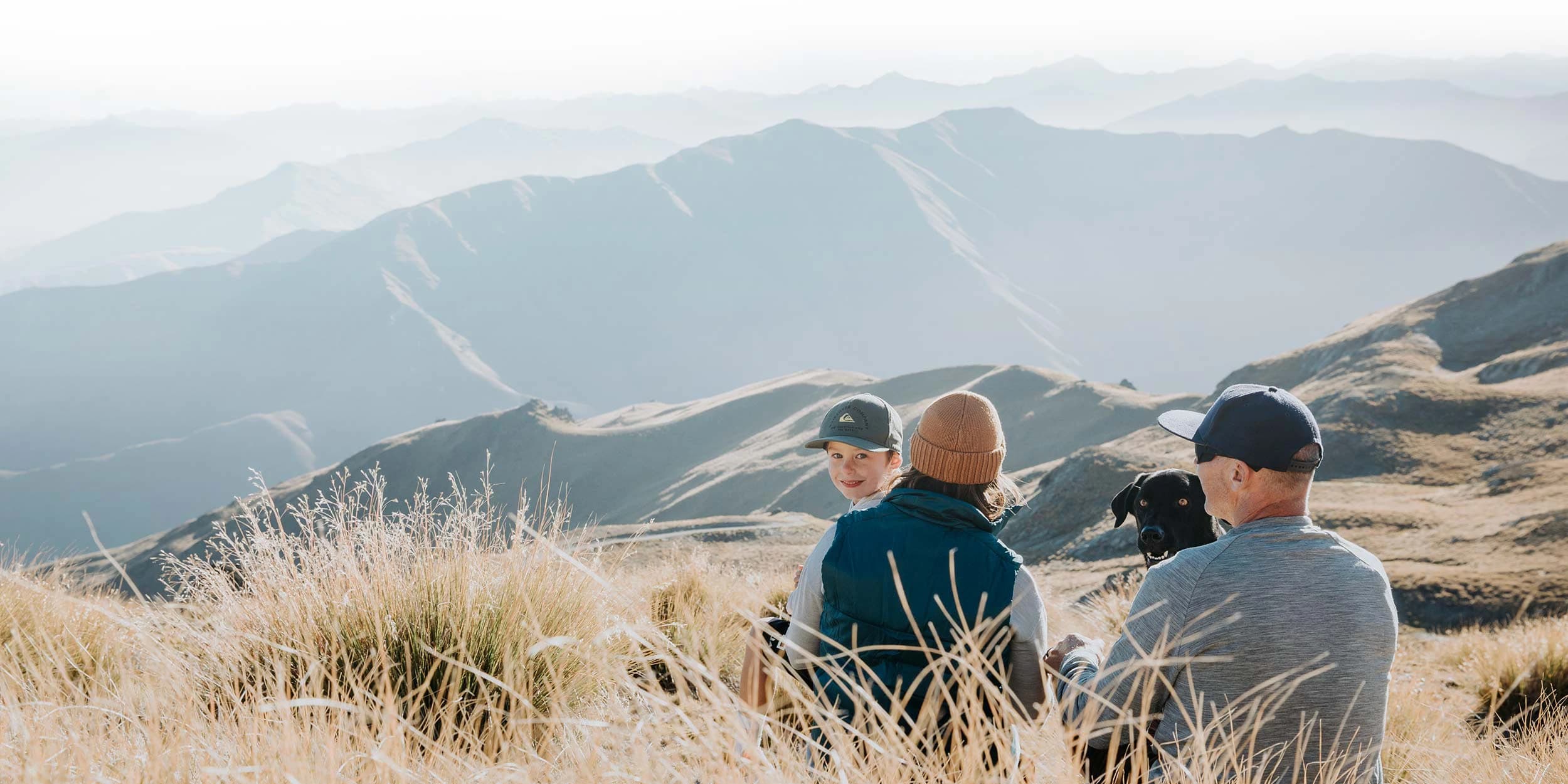 A family of two adults, one child, and a black dog sit amidst tall, golden grass on a mountainous terrain. The background features a panoramic view of softly lit mountain ranges under a clear sky. The child and the dog are facing the camera, while the adults are turned towards each other, enjoying the serene landscape.