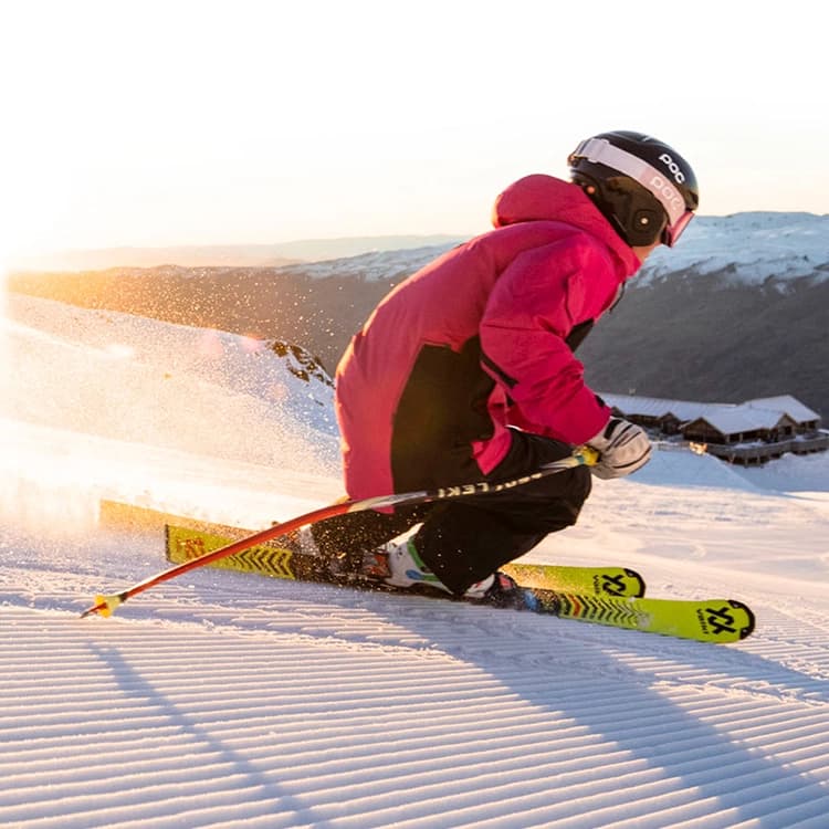 A skier in a bright pink jacket glides down a groomed slope at sunrise, kicking up snow with the mountains in the background and golden light illuminating the scene.