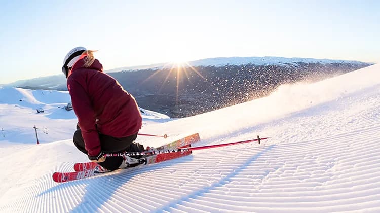 A skier carves through the freshly groomed snow at Cardrona, with the sun rising over the distant mountains, casting a golden light on the slopes as snow sprays up behind them.