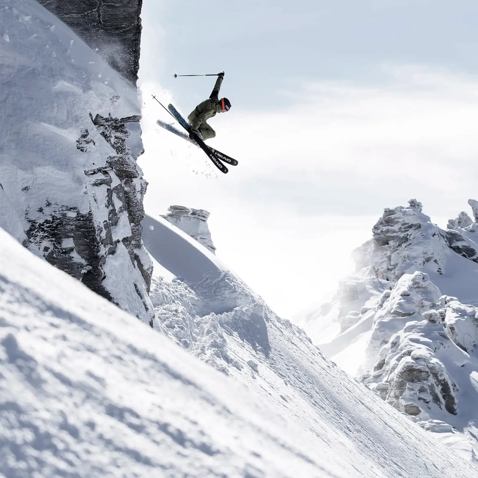 Freestyle skier launching off a steep cliff with snowy rock formations in the background, capturing an adventurous moment in the mountains.