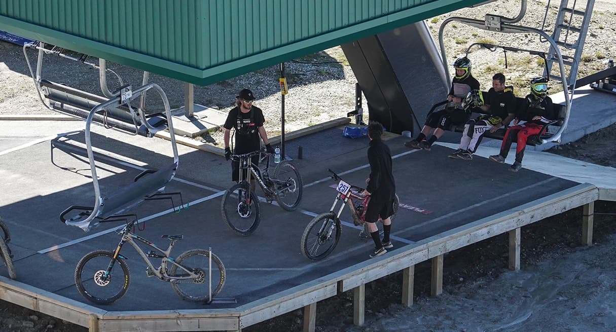 Mountain bikers wait at a chairlift station at Cardrona Alpine Resort, bikes ready on the platform. Riders rest, preparing for their next ride.