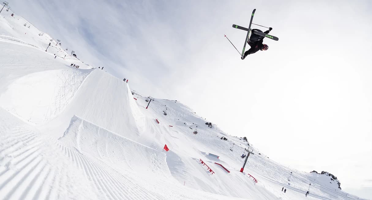 A skier performs an aerial trick high above a terrain park at Cardrona Alpine Resort. Below, the snow-covered park features jumps, rails, and red flags.