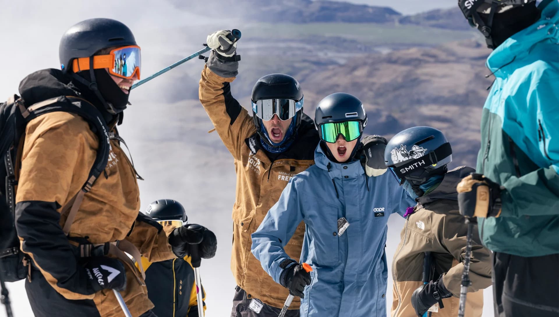 A group of skiers on a snowy mountain. In the foreground, three skiers express excitement and energy: one skier is shouting and pointing skyward, another is cheering, and a third is gesturing playfully towards the camera. The background shows a wide, expansive view of the mountain landscape under a clear sky.