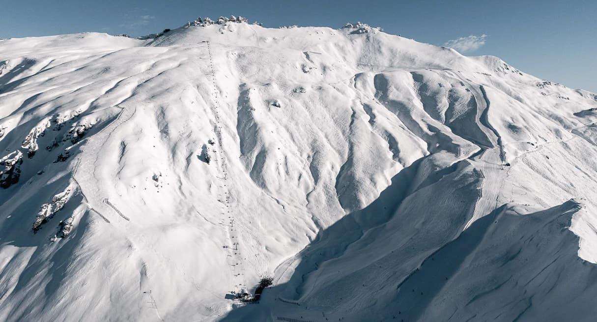 An aerial view of the snow-covered Saddle Basin at Treble Cone Ski Field, showing steep, rugged terrain and a lift line stretching up the mountain under a clear sky.