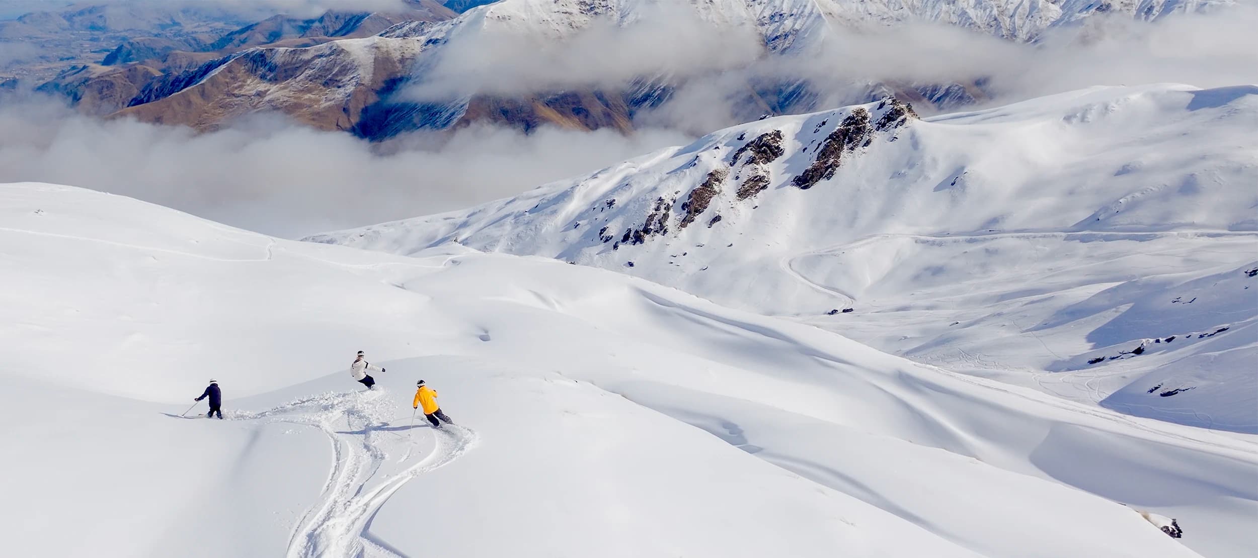 Three skiers glide down the snowy slopes of Soho Basin at Cardrona Alpine Resort, surrounded by vast, untouched powder and majestic mountain peaks. The scene is set against a backdrop of cloud-covered ridges and snow-capped peaks, offering a pristine and expansive winter landscape.