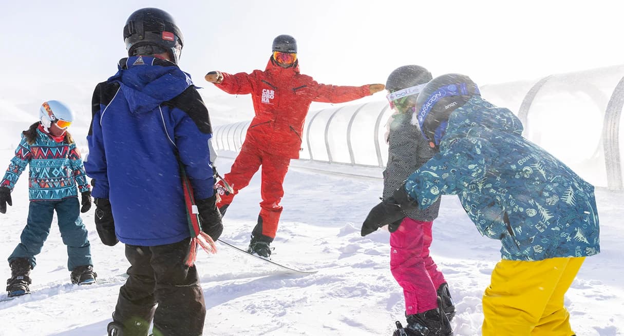 A ski instructor in a red suit teaches a group of young children at Cardrona Alpine Resort. The kids, dressed in colorful winter gear, listen attentively as they stand on a snowy slope near a covered magic carpet lift.