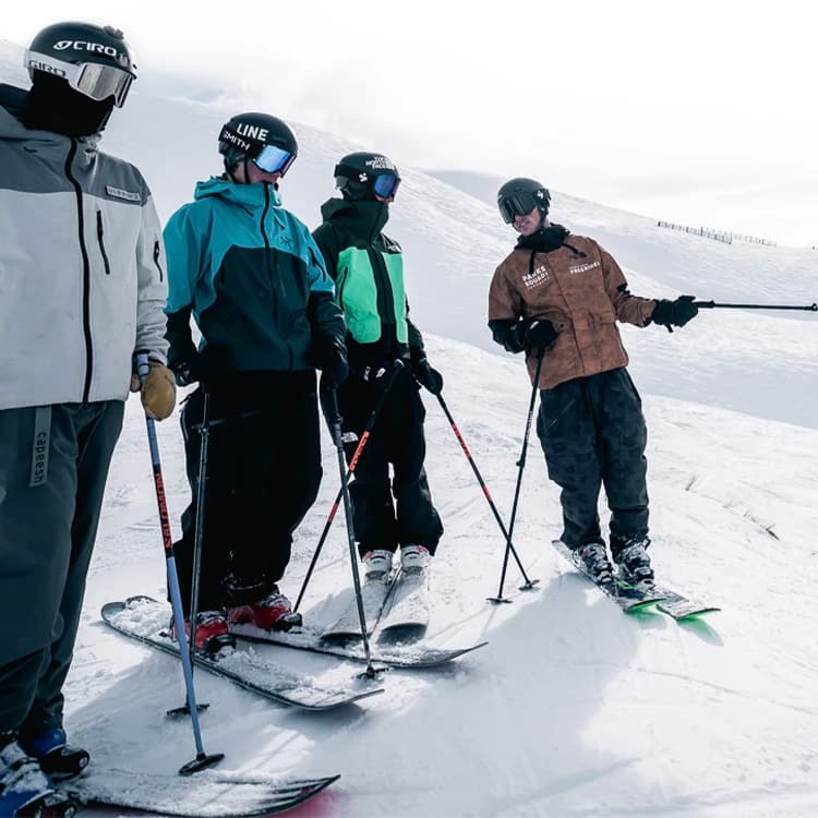 Four skiers on a snow-covered slope, dressed in winter ski gear including helmets and goggles. The skiers are equipped with poles and are standing on skis.