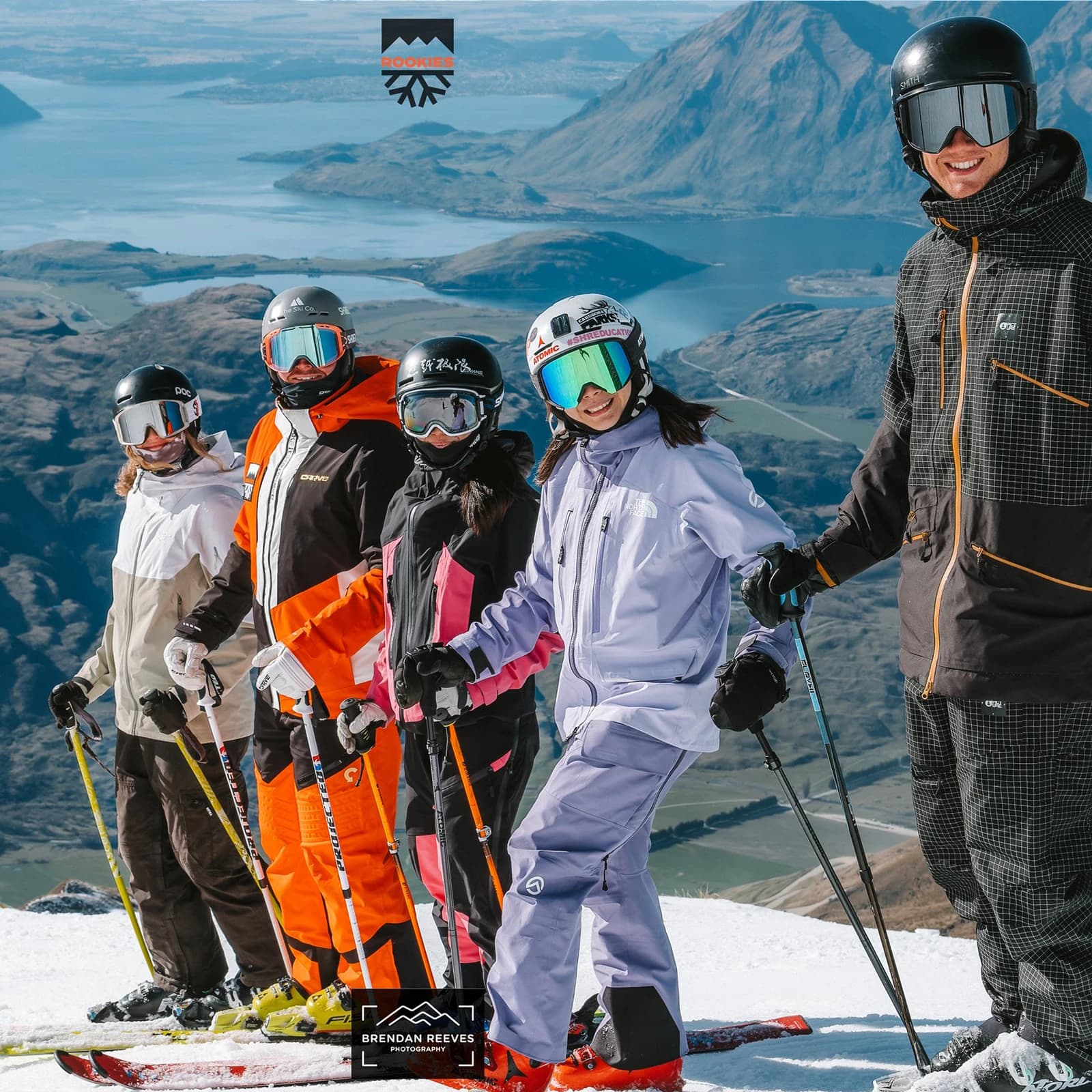 A group of five skiers, dressed in colourful ski gear, pose on a snowy slope with their ski poles, smiling towards the camera. Behind them, a view of a large blue lake, green valleys, and rugged mountains stretches into the distance. The photo features a logo above the group and credits to the photographer, Brendan Reeves, at the bottom.