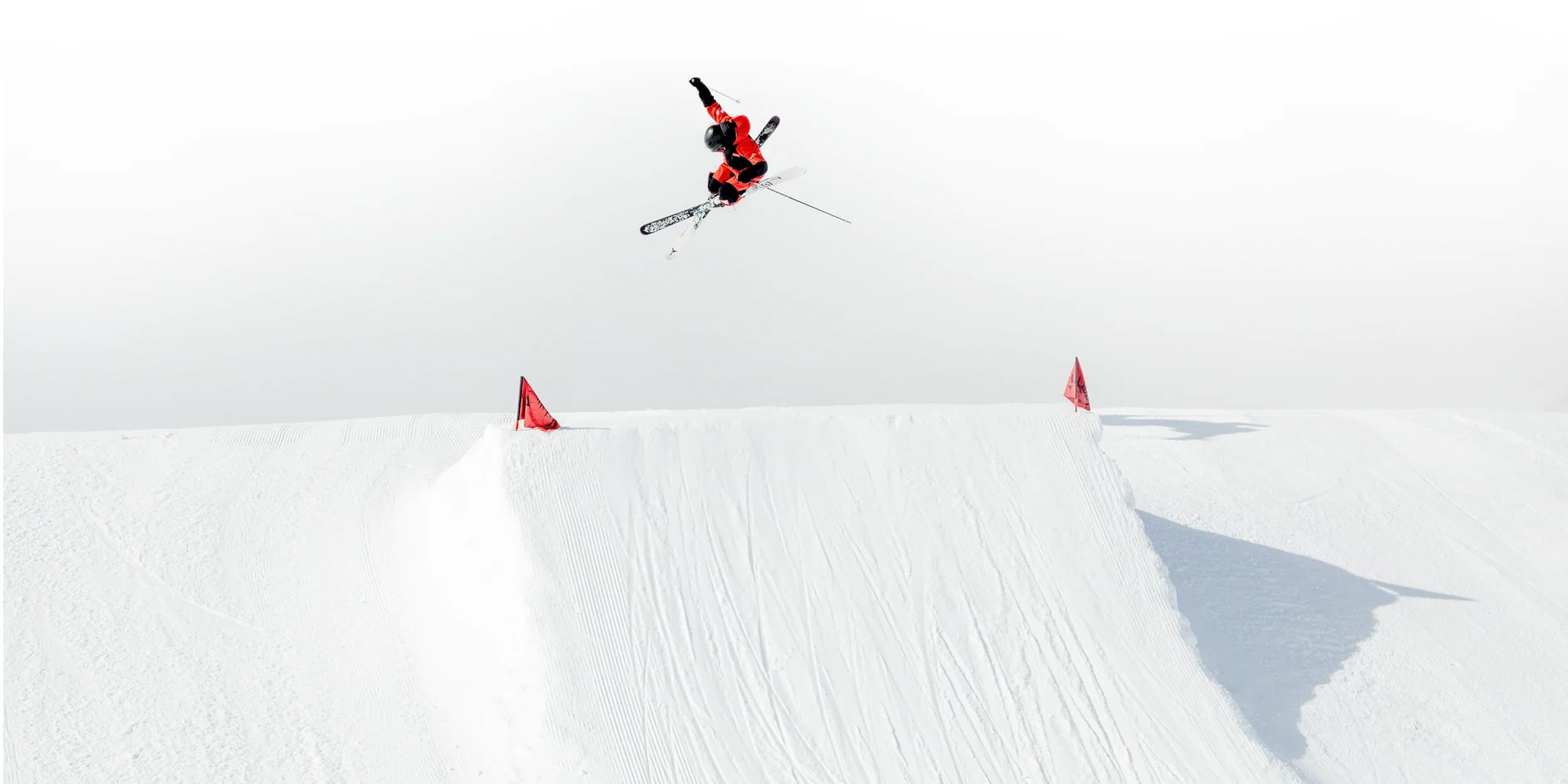 A skier performs a mid-air trick over a snowy slope, with red markers highlighting the edges of the jump, set against a bright, overcast sky.