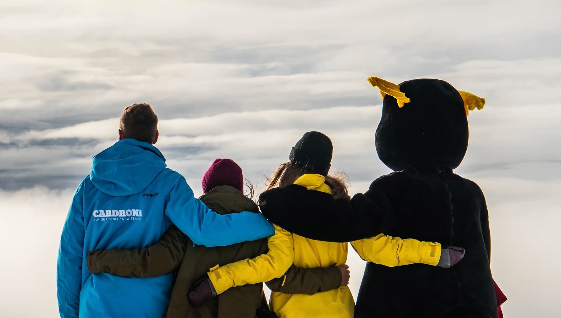 Three staff members and a mascot hold each other and look out at a stunning mountainous view through the clouds.