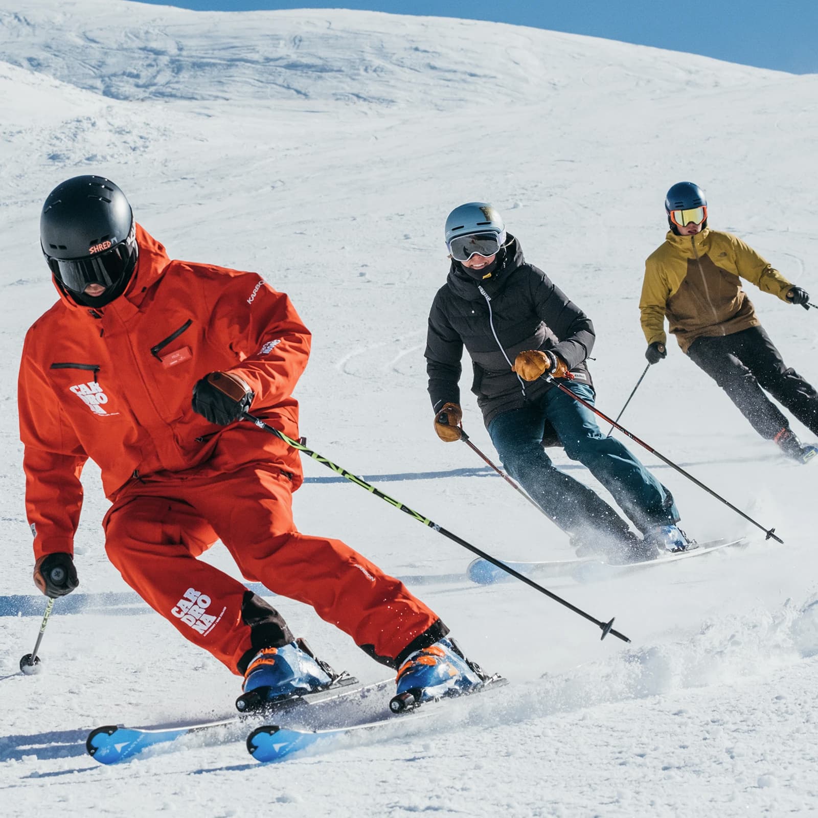 Three skiers expertly navigating a snowy slope under clear blue skies. The skier at the front, dressed in a vibrant red ski suit branded 'Cardrona', is leading the other two in a lesson at Cardrona Alpine Resort.