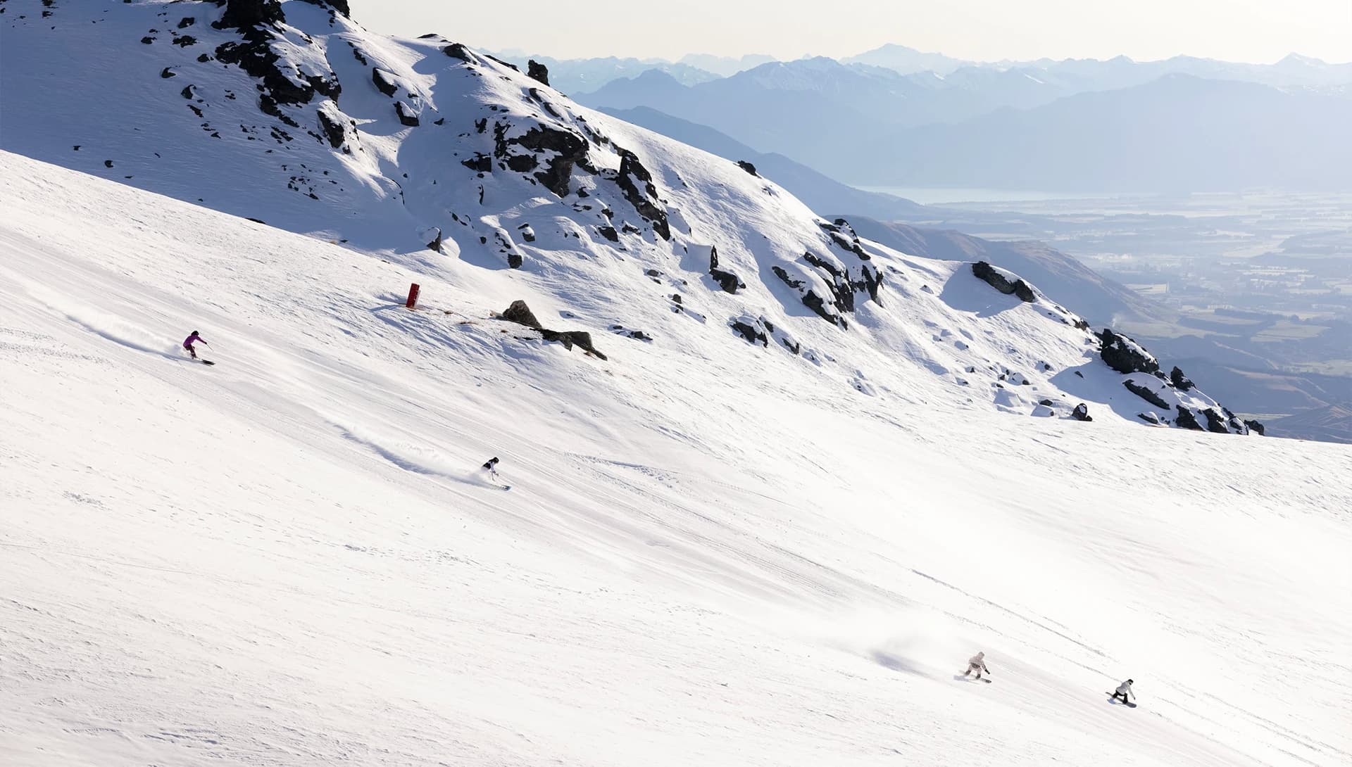 The image shows two skiers and two snowboarders carving down a snow-covered slope at Cardrona Alpine Resort, with rocky outcrops nearby. In the distance, a stunning view of Lake Wanaka and layers of mountain ranges stretch across the horizon, creating a breathtaking alpine backdrop.