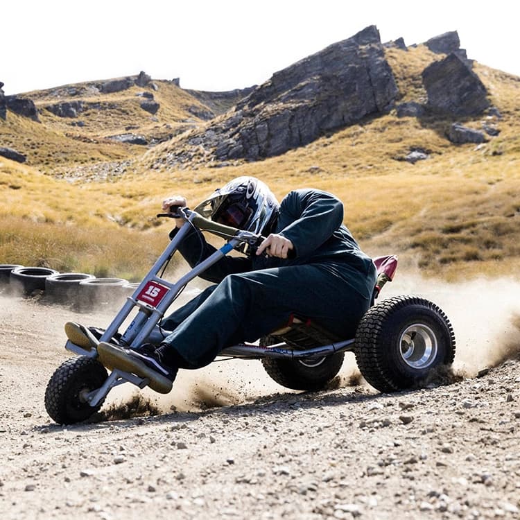 The image captures an intense moment of gravity carting at Cardrona Alpine Resort. A rider in a dark green outfit and helmet leans into a sharp turn on a dirt track, kicking up dust and gravel as they navigate the course. The rugged, mountainous terrain with grassy hills and scattered rocks provides a dramatic backdrop to the action. The rider’s focused posture and the dirt flying from the wheels highlight the adrenaline-pumping excitement of mountain carting at the resort.